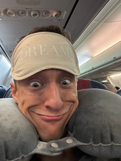 A man wearing a tan sleep mask with "DREAM" printed on it smiles at the camera while on an airplane. He is using a gray neck pillow for comfort. Airplane fixtures and seats are visible in the background. The image is rotated, so the subject appears sideways.