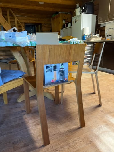 The image shows a wooden dining table with a light-colored wood grain, placed on a light wood-patterned floor. The table has a unique, curved design, with its legs extending upwards and forming part of the tabletop structure. On the table, there is a product box for a Ninja Foodi blender, featuring an image of the appliance and its accessories. To the left of the table, part of a white kitchen appliance, possibly a refrigerator or a large food processor, is visible. Beyond that, a wooden staircase leads upwards, and the walls are made of rough-hewn wood logs, suggesting a rustic or cabin-like setting. A blue cloth is draped over a chair or stool under the table. The lighting suggests it is daytime, with shadows indicating light sources from the right and above. The scene appears to be indoors, likely in a kitchen or dining area of a residential property in Stīveri, Latvia, given the provided location context. There are no people visible in the image, and the overall mood is calm and undisturbed. Visible text includes "Ninja Foodi" on the product box.