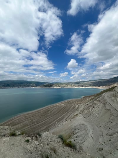 The image presents an aerial view of a landscape. On the right side, there is a rocky, mountainous terrain with sparse vegetation. A body of water, resembling a lake or reservoir, extends lengthwise along the image, and the sky above is filled with blue patches and fluffy white clouds. The location is Chirkey, Russia.