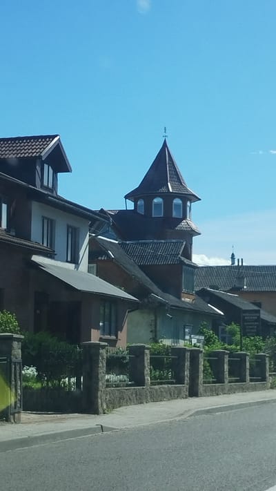 The image shows a street scene in Deliatyn, Ukraine, with a row of buildings along a street. The architecture includes a building with a distinctive tower, topped with what appears to be a weather vane. Other buildings exhibit various rooflines and window arrangements. The street is asphalted, and there are fences or walls separating the properties from the road, some with greenery planted within the stone sections. The sky is blue. The image is oriented on its side.