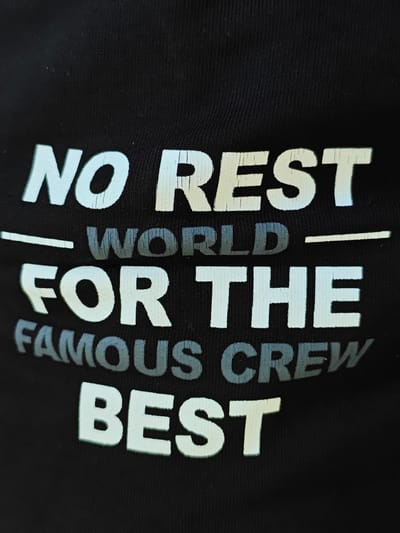 The image shows a close-up of a black t-shirt with white text printed on it. The text reads "NO REST WORLD FOR THE FAMOUS CREW BEST". The text appears to be slightly faded or worn, with some cracks in the white lettering. The t-shirt is made of a ribbed fabric. There are no people or any other objects visible in the image. The location of Crasnogorca, Moldova is not depicted in the image.