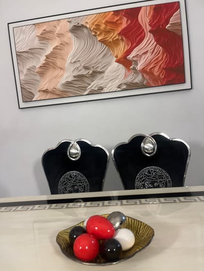 An indoor scene showing a framed abstract art print on a white wall, two black chairs with silver accents, and a gold-colored bowl containing decorative balls. The art print features layered, wavy patterns in shades of red, orange, and beige. The chairs have a distinctive design with silver ornamentation. The decorative balls in the gold bowl are colored red, white, black, and silver.
There is no explicit information regarding the location in the city of Mopo-Akinlade, Nigeria.