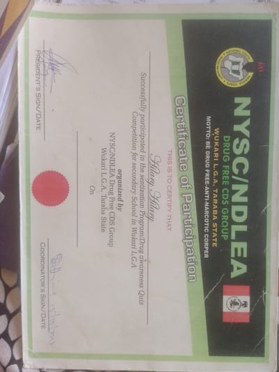 This is a certificate of participation from the NYSC/NDLEA Drug Free CDS Group, Wukari L.G.A., Taraba State, Nigeria. The certificate acknowledges the successful participation in a sensitization program/drug awareness quiz for secondary schools in Wukari L.G.A. The certificate is addressed to "Hilary Hilary." It includes signatures and dates from the President and Coordinator. The background features the NYSC logo and the Nigerian flag. The image is taken indoors, likely in an office or school setting, given the formal nature of the document. The lighting suggests ambient room lighting.