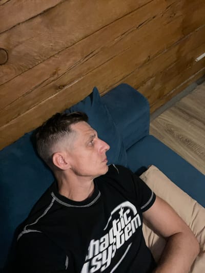 A man in a black t-shirt with white stitching lies on a blue sofa. The t-shirt has a white graphic that reads "Hattic System". He is looking to the right. The background consists of a wooden wall and wooden floor. The man appears to be in a residential setting, possibly a living room, within the city of Polevoi, Russia.