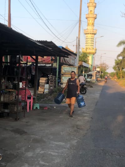 In the city of Besuki, Indonesia, a man wearing a dark tank top and orange shorts walks on a paved road carrying two large blue water jugs. He is walking away from a roadside stall with a dark corrugated metal roof. Behind him, a tall, decorative minaret of a mosque rises into the sky. The scene depicts a typical street view in a small town, with a mixture of commercial and residential structures. The lighting suggests it is either early morning or late afternoon.