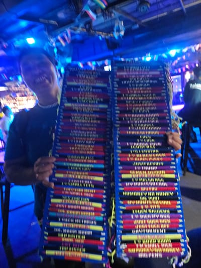In a dimly lit setting illuminated by vibrant blue and purple lights, a person is holding two colorful, rectangular objects that appear to be stacks of bracelets or similar decorative items. The objects are composed of many vertical strands of yarn or string, each in a different color, including red, yellow, purple, and black. White strings are tied at the top of each stack, forming a decorative fringe.

The person holding these items has their face visible in the lower part of the frame. They are smiling and looking slightly upwards. Their clothing is dark. Behind them, more of the colorful stacked items are visible, along with blurred figures and more of the ambient lighting. The scene appears to be indoors, possibly at a market or festival, given the bright lights and the nature of the items being displayed.

Visible text on the items includes repeated phrases in what appears to be English. Some legible words are "PUTIN MIGHTY", "NON-MONEY", "NON-MONEY", and "FIGHTER". The text is printed in yellow and white against the colored strands. The overall impression is of a lively and colorful environment, possibly a tourist-oriented setting. The time of day is likely evening or night due to the artificial lighting.