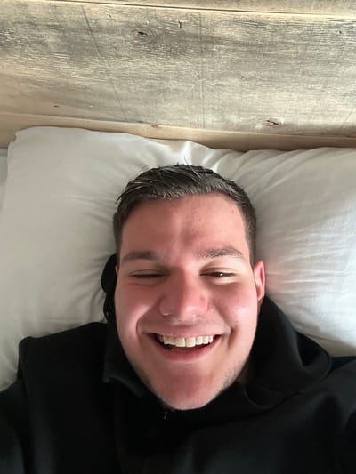 A man is lying on a white pillow next to a wooden wall. He is smiling and wearing a black top. There is no indication of the location being Pamporovo, Bulgaria.