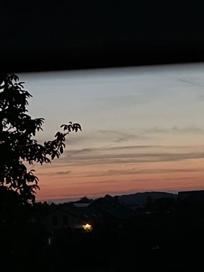 The image captures a twilight scene in Mondercange, Luxembourg, with the sky displaying a gradient of pale blue and soft orange hues. Silhouetted against this sky are the branches of a tree on the left and a distant treeline and rolling hills. Below the sky, a collection of rooftops and buildings is visible in shadow, with a single illuminated window in one of the structures casting a warm glow. The overall impression is one of a peaceful evening transitioning into night.