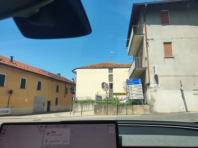 The image is a street view in Borghetto, Italy, captured from a vehicle. In the foreground, a car's dashboard displays navigation information. The scene unfolds to reveal a street with buildings on either side. To the left, a long, two-story yellow building with red tile roofing and green shutters is visible. A white construction barrier is present near the building.

To the right, a taller, light gray building with multiple balconies and red-shuttered windows dominates the view. A series of blue directional signs are mounted on a pole in front of this building, indicating "AMENO," "ARMENO," "municipio," "posta," "scuole," "biblioteca," and "farmacia." A green pharmacy cross sign with a digital display showing "21.6" is also visible further down the street.

The sky is clear and blue, suggesting a sunny day. The road surface is asphalt, and cobblestones are visible on the sidewalk area. The environment appears to be a moderately urban setting, possibly a small town or village.