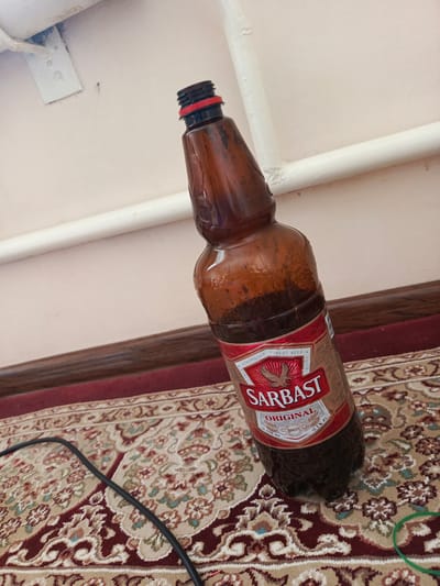 An amber-colored plastic bottle of "SARBAST ORIGINAL" beer, 1.5 liters with 10.5% alcohol by volume, is resting on a red and beige patterned carpet. The bottle is tilted at an angle, with the cap a black and red plastic screw-top. The label is red and white, featuring an eagle and text in what appears to be Cyrillic and Latin script, indicating it is from Uzbekistan. The bottle has condensation on its surface. In the background, a white pipe runs horizontally along a cream-colored wall. A black cable snakes across the carpet, partially obscuring it. A green rubber band is visible on the carpet near the bottom right of the frame. The scene appears to be indoors, in a residential setting. The specific location within the city of Nukus, Uzbekistan, cannot be determined from the provided image.
