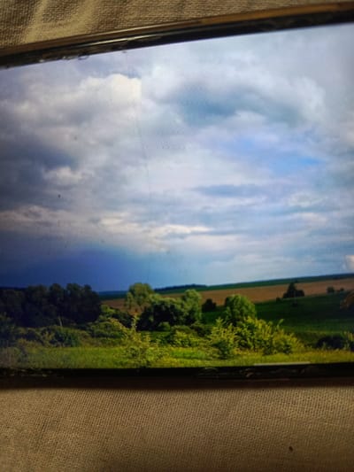 The image depicts a landscape scene viewed on a screen, with the screen edges and surrounding textured material visible. The landscape itself features a dramatic sky with dark, ominous clouds in the left half transitioning to lighter, brighter clouds and blue sky on the right. Below the sky is a horizon line, separating it from a rolling green landscape. In the foreground, lush green trees and bushes are visible, some of which appear dense and others more sparse. The middle ground shows fields, with one appearing as a cultivated brown field and others as green pasture. The overall impression is of a rural or semi-rural environment, possibly a field or parkland, under a dynamic sky. There are no people or identifiable structures present in the landscape. The image is likely a photograph taken of a screen displaying a landscape, indicated by the horizontal lines across the image which are characteristic of screen display artifacts. The surrounding context suggests the image is displayed on a device placed on a coarse, woven surface. The specific location of Svoboda, Russia, is not visually identifiable from the content of the image itself.