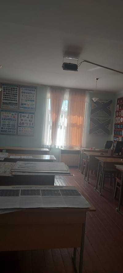 The image depicts the interior of a classroom, likely in Tetiiv, Ukraine, characterized by rows of wooden desks and a projector mounted on the ceiling. The walls are adorned with various educational charts, predominantly featuring road signs and instructional diagrams. A large window with sheer, peach-colored curtains dominates one wall, allowing diffused sunlight to illuminate the room. On the desks, stacks of papers and posters are visible, some appearing to be study materials. One desk is equipped with a computer monitor, suggesting the room may also be used for computer-based learning. The floor is made of dark, wooden planks, and the overall atmosphere is one of a functional, albeit somewhat dated, educational space. The lighting is soft, with shadows cast by the desks and projector. No individuals are present in the scene.