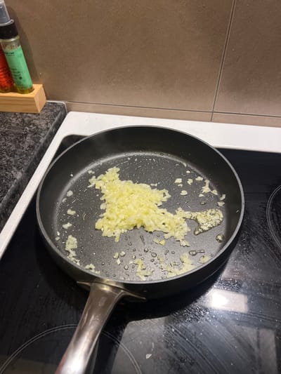 A close-up, high-angle image depicts a black non-stick frying pan situated on a modern black ceramic cooktop in a kitchen. Inside the pan, a substantial amount of finely diced white ingredients, consistent with chopped onions or garlic, is visible, glistening with small droplets of oil. This suggests the ingredients are being, or are about to be, sautéed. The frying pan has a distinctive metallic silver handle extending towards the right side of the frame.
To the immediate left of the cooktop, a light brown tiled wall serves as a backsplash. Below this tiled wall and extending towards the foreground is a dark, speckled countertop. On this countertop, positioned on the far left, a small wooden block holds several test-tube-like containers. One of these containers, which is green, shows partially visible white text, including the word "PURE" and what appears to be "FINE" or "FIE", indicating a spice or seasoning.
The scene captures a moment of food preparation within a contemporary kitchen setting. The lighting is consistent with indoor artificial illumination. This activity takes place in a kitchen located in Chêne-Bourg, Switzerland.