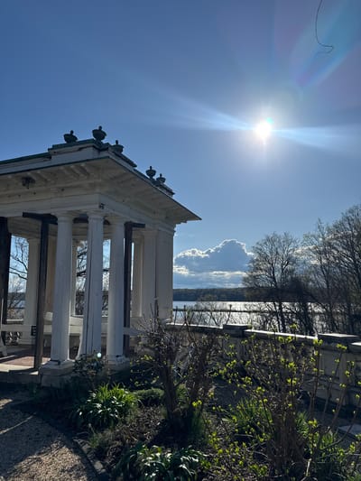 The image shows an outdoor scene featuring a white gazebo-like structure with classical columns, situated near foliage and trees. The sky is clear blue, with the sun shining brightly, creating lens flare in the upper left. A cloud is visible in the background. The gazebo is likely located in a park or garden setting. The image was taken in Town of Red Hook, United States of America.