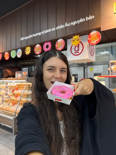 A young woman with long, dark, wavy hair is holding a white cardboard container with a pink frosted donut covered in sprinkles. She is looking at the camera with a slight smile, and her mouth is open as if about to take a bite of the donut. She is wearing a black garment. In the background, there is a display case filled with baked goods, and signs are visible on the wall above the display. One sign has Vietnamese writing and colorful circular decorations, possibly indicating sales or promotions. The overall setting appears to be a bakery or a food establishment. The question mentions the location as Phu Thuy, Vietnam, which is relevant context for the signage.
