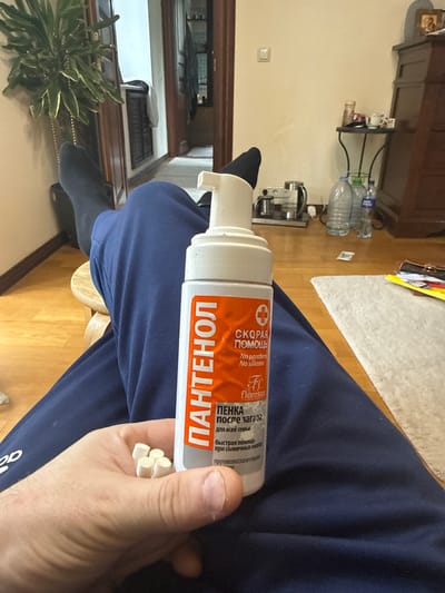 A hand holds a bottle of "Panthenol" foam, designed for after-sun care, with Russian text on the label, along with some white sticks. The person is wearing blue sweatpants and sitting on a wooden chair. The background is blurred, showing a room interior with a wooden cabinet, bottles, and a plant. The location is Lesnoi Gorodok, Russia.