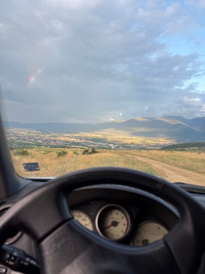 This media file shows a view from the driver's seat of a vehicle, looking out at a rural landscape. The steering wheel is visible on the right side of the frame, with two gauges partially obscured behind it. The landscape outside features rolling hills covered in dry grass, with a small town nestled in a valley below. In the distance, large mountains rise, their peaks shrouded in clouds. The sky is partly cloudy, with patches of blue peeking through. A faint rainbow is visible in the lower left portion of the image, suggesting recent rain or mist. The overall impression is of a scenic drive through a countryside setting, possibly in the late afternoon, given the quality of light. There is no visible text in the image.