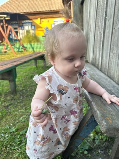 A young Caucasian female child, approximately one year old, stands outdoors. She has blonde hair tied in a ponytail with an orange band and blue eyes. She is wearing a white dress adorned with a pattern of teddy bears and bows. Her left hand holds a green leaf, and her right hand rests on a weathered wooden bench. The bench appears old and weathered, with traces of blue paint visible on its supports. Behind the child, a rustic wooden fence and parts of a house with a corrugated roof are visible, suggesting a rural or suburban setting. The ground is covered in green grass and some scattered foliage. The lighting indicates it is daytime, likely late afternoon, with warm sunlight casting shadows. The overall scene depicts a quiet moment of a child exploring nature in a possibly rural Russian environment, consistent with the outskirts of a town like Novoyeniseyka, Russia, characterized by simpler wooden structures and natural surroundings.
