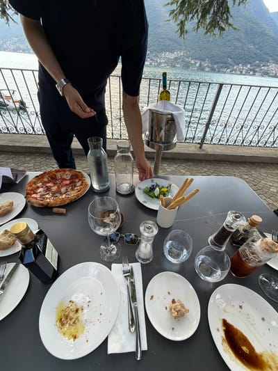 An outdoor dining setting at a restaurant in Pognana Lario, Italy, is depicted. A person, partially visible from the waist down, wearing a dark blue shirt and pants, stands near a table laden with partially eaten food and drinks. The table is set with white plates, silverware, glasses, salt and pepper shakers, a bottle of olive oil, and a pizza. A bottle of white wine rests in an ice bucket in the background. The table overlooks a lake, with the town of Pognana Lario visible on the hillside across the water. The atmosphere suggests a meal in progress or recently concluded.