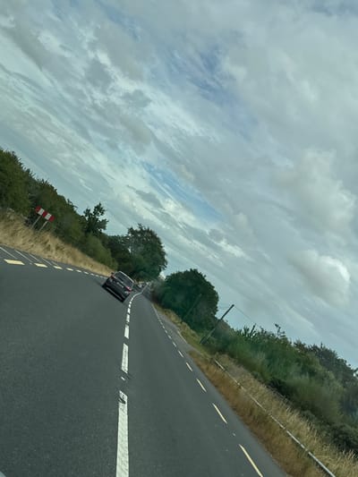 The image shows a view from a vehicle traveling on a rural road in New Ross Municipal District, Ireland. The road is asphalt with white and yellow markings. On the left side of the road, there is a red and white striped sign indicating a hazard or turn. The roadside is bordered by dry grass and dense green bushes and trees. In the distance, other cars can be seen on the road. The sky is cloudy with patches of blue visible. The image is taken from a low angle, suggesting it was captured from within a car.