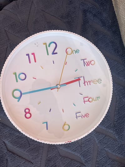 A white circular clock with colorful numbers and words is displayed on a dark blue textured fabric surface. The clock has a scalloped edge and a white frame. The numbers 1 through 12 are represented by colorful digits and written-out words. The hour hand is red, and the minute hand is light blue. The clock appears to be a decorative item, possibly for a child's room. The overall scene is static and does not depict any specific action or interaction. There is no information in the image to suggest a location in Zlataritsa, Bulgaria.
