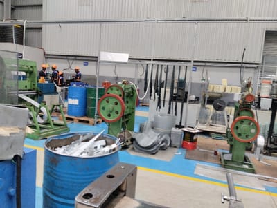 The image depicts the interior of an industrial workshop or factory floor, located in Ngecha Tigoni ward, Kenya. The facility features a blue floor with yellow demarcation lines, and light grey corrugated metal walls, typical of a manufacturing environment.
In the mid-ground, four workers are visible, all wearing orange safety helmets and uniforms that appear to be blue and orange. They are gathered around a large green industrial machine, seemingly operating or overseeing a metal processing task. Metal strips or sheets are seen feeding into or coming out of this machine. Another similar green machine with large red and green wheels stands further to the right.
Several blue and green industrial barrels are present, suggesting storage of liquids like oil, coolants, or other materials. One blue barrel in the foreground is filled with numerous shiny, elongated metal scraps, indicating metal cutting, stamping, or shearing operations. Stacks of processed or raw metal sheets and various other containers and equipment are scattered throughout the workspace.
The overall lighting suggests it is daytime, likely due to artificial industrial lighting within the building. The scene portrays an active work environment focused on metal fabrication. While there are distinct work areas, some equipment and materials appear somewhat arranged and others casually placed. No specific text is clearly legible on any objects or labels.