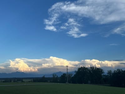 A wide, horizontal, eye-level shot captures a vast expanse of blue sky with dramatic, billowing white clouds, illuminated by what appears to be late afternoon or early evening sunlight. The lower third of the image is dominated by a verdant, grassy field stretching towards a dark, silhouetted tree line. In the distance, hazy blue mountains form a backdrop against the horizon. A single, dark utility pole with wires is positioned slightly off-center to the right in the middle ground, breaking the natural landscape. The scene is likely in or near the Bavarian village of Höslwang, Germany, given the characteristic landscape and clear sky with developing cloud formations. There are no visible people or animals.
