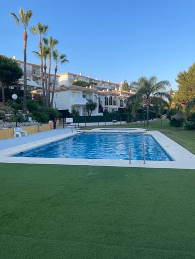 This is a wide, eye-level, outdoor, daytime, full shot of a swimming pool area in Mijas, Spain. In the foreground is a rectangular swimming pool with bright blue water, surrounded by white tiling. Two silver railings rise from the pool for entry and exit. The pool area is bordered by a strip of artificial green grass, and beyond that, more of the artificial turf lawn covers the ground. In the midground, behind the pool, there is a paved walkway with a white bench and a trash can. Behind the walkway, there is a fence and then a hill covered with lush green vegetation and several tall palm trees. In the background, there are several white residential buildings with terracotta tile roofs, characteristic of Mijas architecture. The sky is clear and blue. The image captures a serene and well-maintained recreational space.