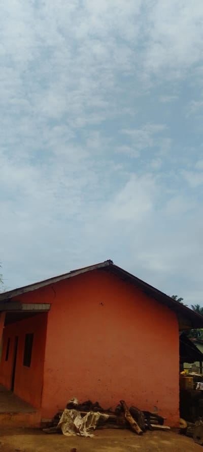 The image captures a side view of an unfinished or weathered orange building under a cloudy sky. The building has a simple, pitched roof with exposed wooden beams along the edge. On the left side of the building, there are two dark rectangular openings that appear to be windows or doors. The exterior wall of the building shows some cracks and a slightly rough texture.

In front of the building, the ground is uneven and dusty, with some scattered debris including pieces of wood, cloth, and possibly other building materials. Some of these materials are piled together, suggesting they might be stored or awaiting use. Further to the right, partially visible behind the main structure, is another darker roofed area or shed, with some yellow and white objects that could be containers. Palm trees are visible in the background, suggesting a tropical or sub-tropical environment.

The sky is filled with a layer of diffused white and grey clouds against a pale blue background, indicating an overcast or partly cloudy day. The lighting suggests it is daytime, possibly late morning or early afternoon, with diffused sunlight. There is no text visible in the image. The overall scene conveys a sense of rural simplicity, possibly in a developing area, given the unfinished look of the building and the surrounding conditions. There are no people visible in the image.