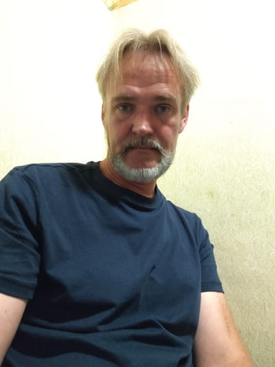The image is a close-up, waist-up shot of a middle-aged man with blond hair and a mustache and beard. He is wearing a dark blue t-shirt. The background appears to be a cream-colored wall with a textured pattern. The lighting is overhead, casting shadows on the man's face and shirt. There are no discernible landmarks or specific architectural features that would indicate a location within the city of Rezekne, Latvia. The image appears to be a selfie taken indoors.