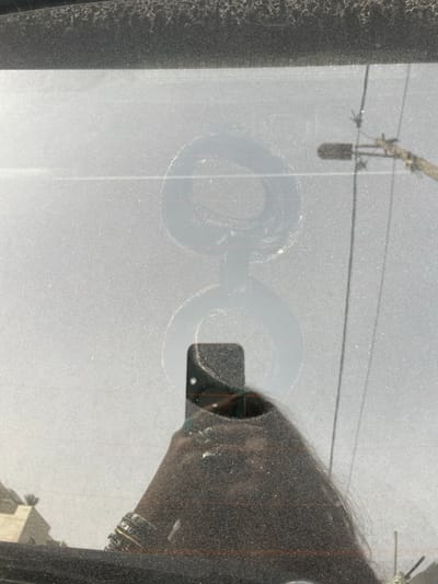 A dusty window reveals a partial reflection of a person holding a cell phone to take the photo. The reflection also shows hair and parts of the car's interior. Outside, a building is partially visible below. The window has two circular smudges or fingerprints in the center. Overhead, power lines and a utility pole are silhouetted against the brighter background. The image has a grainy texture throughout due to the dust on the glass.