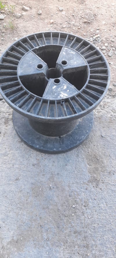 A black plastic cable spool sits on a concrete surface in an outdoor environment. The spool has a large central opening and spokes radiating outwards on its upper surface, with a wider base. The ground around the spool is a mixture of concrete and dirt with small stones. There is no visible damage or wear on the spool, except for some white scuff marks on the inner surfaces of the spokes. The background shows more dirt and stones. The image appears to be taken in Zhasaqan, Kazakhstan.