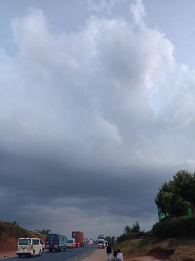 A vertical shot showcases a road bustling with vehicular traffic beneath a sky heavily laden with ominous, dark clouds. The roadside is flanked by greenery and individuals walking. The sky dominates the frame, presenting a gradient from lighter, brighter clouds to dense, storm-like formations. Vehicles line the road, hinting at congestion or slow-moving traffic. The color palette is dominated by grays, greens, and muted earth tones.