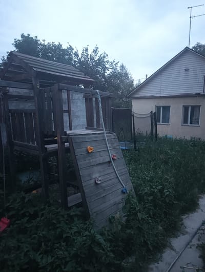 The image depicts an outdoor scene in the city of Rassvet, Kazakhstan. A wooden playground structure, featuring a slide and climbing holds, is situated in the foreground. A rope ladder extends from the structure, partially obscured by dense green foliage. Behind the playground, a modest, light-colored house with a pitched roof and two windows is visible. A trampoline with a black safety net stands in the overgrown backyard. The sky is overcast, suggesting either early morning or late evening. The overall impression is one of a residential yard with a well-used playground, surrounded by abundant vegetation.
