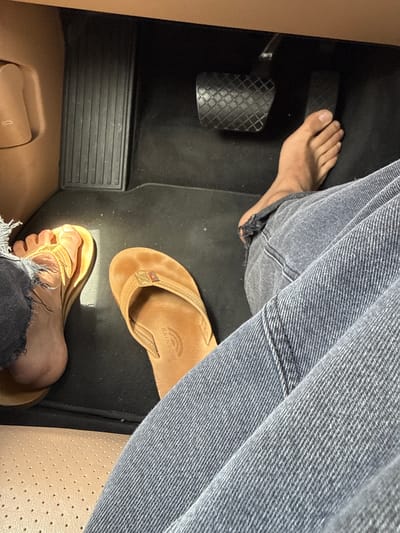 The image shows a person seated in the driver's seat of a vehicle, with one foot wearing a brown flip-flop sandal and the other foot bare, resting on the accelerator pedal. The person is wearing faded blue denim jeans with a distressed hem. The interior of the vehicle is visible, including the pedals, floor mat, and seat upholstery. The make, model, and year of the vehicle cannot be determined. The geographical location cannot be determined from the image.
