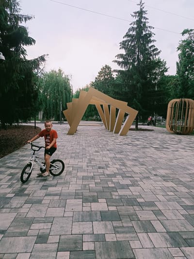 A young boy rides a white bicycle on a paved walkway. He is wearing a red t-shirt with a graphic of Mickey Mouse and black shorts. The walkway is made of square and rectangular grey tiles. In the background, there are trees, an abstract sculpture made of wooden beams, and a circular wooden structure. In the distance, a red ATV with a driver is visible. This scene is in the city of Gubkin, Russia.