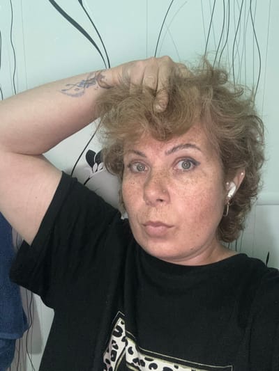 A woman with short, wavy blonde hair and freckles on her face is shown in a close-up, selfie-style shot. She is wearing a black t-shirt and has her left arm raised, with her hand in her hair. She is also wearing white wireless earbuds in her right ear and a small hoop earring in her left ear. A tattoo is visible on her upper left arm. The background consists of a wall with a patterned wallpaper featuring thin, dark vertical lines and some black floral designs. There is a blue towel visible on the left side of the frame. The lighting suggests an indoor setting. There is no indication of the city of Kozmodemyansk, Russia, or any other specific location in this image.