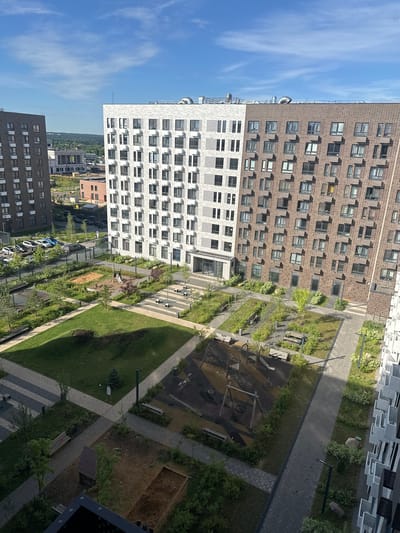 This is an aerial, medium-shot, eye-level photograph of a modern residential complex in Ильинское-Усово, Russia, on a sunny day with a blue sky and scattered clouds. In the foreground, a paved courtyard area features a children's playground with swings and a slide, surrounded by grass and landscaping. Several benches are placed throughout the courtyard. In the midground, a sandpit with a small playhouse is visible. The courtyard is bordered by walkways and residential buildings. The buildings are tall, multi-story structures with numerous windows and balconies, constructed with brick and lighter-colored materials. In the background, other buildings and trees are visible, along with a parking lot with several cars. The overall impression is of a well-maintained, suburban residential area.
