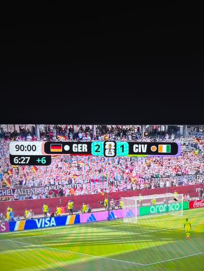 The image depicts a soccer match scoreboard displaying "90:00" and "6:27 +6". The score is "GER 2" against "CIV 1". The crowd in the background is filled with spectators, many holding flags and banners. There are also several people in the foreground, including what appear to be security personnel or team staff. The scene takes place on a soccer field with a goal net visible. Sponsorship logos such as "VISA" and "aranco" are also present. The presence of the German flag associated with "GER" strongly suggests the match is taking place in Germany. Given the specific mention of "Wettstetten, Germany" in the prompt, it is highly probable that this image was taken during a sporting event held in that location.