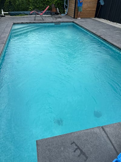This media file shows an aerial view of a rectangular swimming pool filled with clear turquoise water. The pool is surrounded by a concrete deck. To the left of the pool, there is a wooden deck with a person standing on it wearing a blue t-shirt and green shorts. Next to the wooden deck, there is lush green foliage. On the right side of the pool, a dark grey concrete area is visible, with some faint markings that resemble a rake. A bright blue object, possibly a pool float or toy, is partially visible in the top right corner. The water has ripples, suggesting it is either undisturbed or experiencing a light breeze. The scene appears to be outdoors, likely during daylight hours, given the brightness and reflections on the water. The overall impression is a serene and inviting outdoor setting, possibly a backyard in Mississauga, Canada.