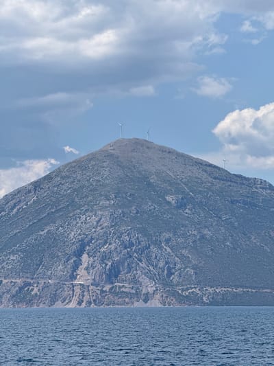 This media file is a photograph of a mountainous coastline with the sea on the right. The image is oriented vertically, so the mountain appears to be on the left and the sea on the right. The mountain is rugged and covered in sparse vegetation. There are three wind turbines visible on the slopes of the mountain, suggesting a renewable energy installation. The sky is blue with scattered white clouds. The sea is a deep blue and appears somewhat choppy, indicating a breezy day. The scene is likely set during daylight hours, given the bright sky. No people are visible in the image. The location is specified as Patras, Greece, which aligns with the mountainous coastal landscape. The overall mood is serene and natural, with a touch of modern industrial presence from the wind turbines. No visible text is present.