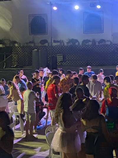 In Blanca, Spain, a crowd of people is gathered outdoors at night, illuminated by blue and white stage lighting. The scene appears to be a social event, possibly a party or concert, as evidenced by the presence of a stage setup with lighting equipment. Many individuals are seen standing and interacting, with some holding drinks and others looking towards a central area, suggesting a focal point of activity. The environment features a building with distinctive arched windows with barred fronts in the background, and a textured lattice-like fence or railing. The ground is paved, and several chairs are visible, indicating a seating area. The attire of the attendees is varied, ranging from casual wear to what might be considered costumes, such as a person dressed in a red outfit with blue overalls, resembling a character. The overall impression is one of a lively, communal gathering under artificial lights.