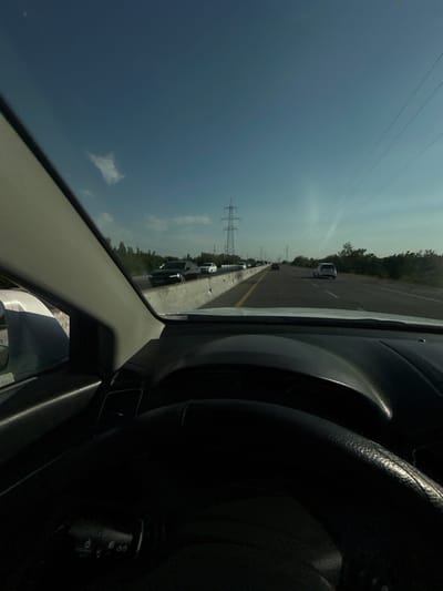 The image depicts a view from inside a vehicle, looking out at a highway. The perspective is from the driver's seat, as evidenced by the steering wheel and dashboard elements visible on the right side of the frame. The highway appears to be congested, with several cars lined up ahead. Trees and utility poles are visible along the roadside, suggesting a rural or semi-rural setting. The sky is a light blue with some clouds, indicating it is daytime, likely late morning or afternoon. The overall impression is of being stuck in traffic on a highway. There is no visible text in the image.