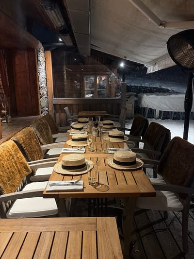 This image captures an empty outdoor dining area, identified as a restaurant terrace in La Clusaz, France, during an evening snowfall. The scene features a long, rectangular wooden table in the center, meticulously set for twelve guests. Each place setting includes a light-colored straw boater hat with a black band, inverted on the table, a white folded napkin with a knife and fork, and a clear drinking glass. Centrally, the table holds a salt and pepper shaker set and a glass bottle.

The chairs along the table are light-toned with white cushions, and many are draped with shaggy brown or amber-colored faux fur throws over their backs. The dining area is covered by an overhead awning structure from which several elongated infrared strip heaters are mounted. A tall, dark, freestanding patio heater with a circular protective grill is positioned on the far right side of the image.

To the left, a rustic wooden wall with stone elements and a prominent horizontal wooden beam defines the interior edge. A transparent barrier separates the covered dining space from the exterior environment. Beyond this barrier, snow is actively falling, evidenced by blurred white streaks against the darker background. The ground outside is covered in snow, and a large white tarp conceals an unidentified object or structure in the middle distance. Distant wooden chalet-style buildings with illuminated windows are visible through the falling snow. No individuals are present in the scene.