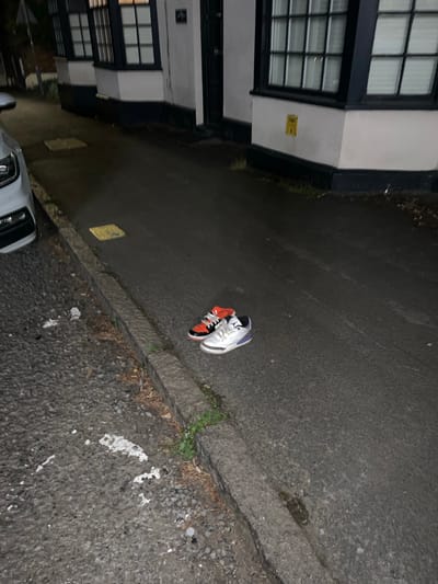 The image shows a nighttime scene in Lane End, United Kingdom. A white building with black-framed windows is visible on the left. On the pavement, there are two sneakers, one orange and white, and the other white and grey. A portion of a white car is visible in the lower left corner of the image. The pavement is made of dark asphalt and there are some patches of grass and debris along the edges.