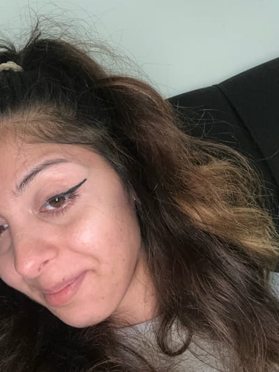 A close-up, low-angle, indoor shot focuses on the upper body and face of a young woman with medium-toned skin, brown hair with blonde highlights, and a grey shirt. She has her hair pulled up with a light-colored elastic band and some loose strands framing her face. She is wearing winged eyeliner. Her left eye is open, and she is looking downwards and to her left. Her mouth is slightly open, and she appears to be smiling or about to speak. The background is blurred, showing a light-colored wall and a dark garment, possibly a jacket or sweater. There is no indication of the city of Augsburg, Germany, in this image.