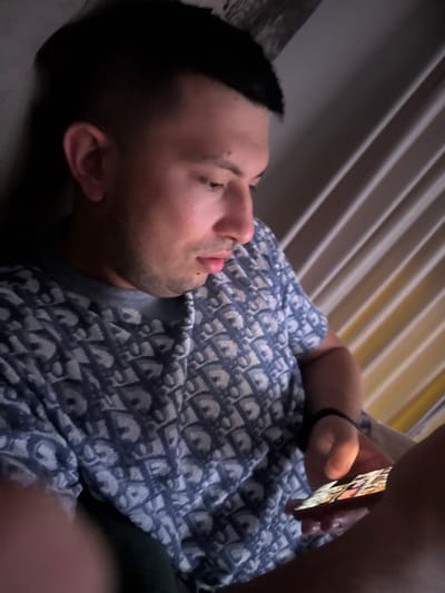 A young man of apparent Bulgarian ethnicity, with short dark hair and a light complexion, is seen from the chest up, reclining and looking at a smartphone in his hand. He is wearing a blue t-shirt with a repeating "Dior" pattern. The background consists of what appears to be blinds or a textured wall. The lighting suggests an indoor setting, possibly at night, with the smartphone screen illuminating his face. The image was taken in Slanchevo, Bulgaria.