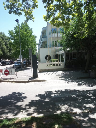 This media file captures an outdoor scene on a sunny day, likely in the afternoon given the strong shadows cast by the trees. The setting appears to be a street in Banyoles, Spain, with a modern building, possibly a hotel or office, as the backdrop. The building has a white facade with multiple stories, featuring balconies with glass railings and large windows. Several tall, slender poles stand in front of the building, adding an architectural element.

The foreground is dominated by a street and sidewalk area. A bicycle is visible, parked near a temporary barrier made of metal fencing and red and white circular signs, which indicate "no entry" or prohibition. Several cars are parked along the side of the road, including a dark blue sedan. Lush green trees with dense foliage are prominent throughout the scene, providing shade and framing the view. One large tree on the right side of the frame has a thick trunk and branches that extend towards the viewer.

The ground surface consists of asphalt on the street and paved stone or tile on the sidewalk. Shadows from the trees create dappled patterns on the pavement, suggesting a bright, clear sky overhead. There is no visible text that is legible or contextually significant in the image. The overall impression is one of a tranquil, urban environment on a pleasant day. No people are clearly visible in the image.