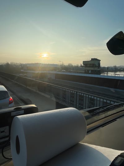 The image depicts a panoramic view from the interior of a vehicle, looking through its windshield during either sunrise or sunset, indicated by the low sun casting warm light and lens flare across the scene. A large, white roll of paper towels is positioned prominently on the vehicle's dashboard in the foreground, partially unrolled onto a dark surface.
In the midground, a multi-lane road or bridge is visible, with several passenger cars present. A dark metal railing runs along the edge of this structure.
The background reveals a large infrastructure complex associated with the Iffezheim Lock (Schleuse Iffezheim) on the Rhine River in Iffezheim, Germany. To the left, a modern, multi-story white building with a flat roof and extensive glazing is discernible, possibly an administrative or customs facility. To the right, a distinct, multi-level control tower with industrial features overlooks a body of water, identifiable as the Rhine River. Beyond the structures, a line of deciduous trees forms the horizon against a clear, pale sky. No individuals are clearly identifiable.