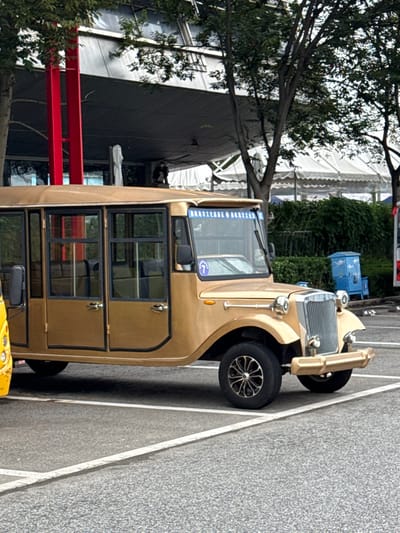 A gold-colored, retro-styled electric tour bus, designed with classic automobile aesthetics, is stationary in a paved parking area within the Shinan District of Qingdao, China. The vehicle exhibits a chrome-like front grille, circular headlights, a gold-colored bumper, and multiple framed glass windows along its side. A blue banner across the windshield identifies it with the 'Qingdao Olympic Sailing Culture Tourism Area,' and a blue circle with the number '7' is affixed to the passenger side windshield. The ground is dark gray pavement marked with white parking lines. In the background, deciduous trees partially obscure a modern building featuring a dark gray facade and prominent red structural columns, as well as a large white event tent. A blue waste receptacle is positioned near green shrubbery. No active human presence is distinctly identifiable.